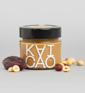 jar of hazelnut and dates spread with black lid surrounded by hazelnuts and a date all on light grey background